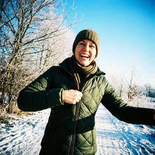 Eine Frau lacht in die Kamera, während sie sich eine warme Jacke anzieht, im Hintergrund eine winterliche, aber sonnige Landschaft.