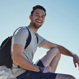 Ein Mann in einem grauen T-Shirt, Shorts und einem Rucksack sitzt im Freien auf einem Felsen und lächelt in die Kamera unter einem klaren blauen Himmel.