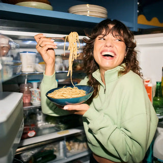 Eine Frau steht lächelnd vor einem offenen Kühlschrank und hält mit einer Gabel eine Schüssel Spaghetti in der Hand.