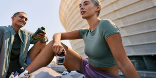 Zwei Personen in Sportkleidung sitzen im Freien in der Nähe eines modernen Gebäudes, halten Nahrungsergänzungsmittelflaschen und Getränke in den Händen und wirken nach dem Training entspannt.