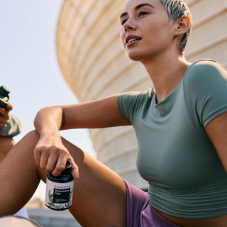 Eine Person in Sportkleidung sitzt im Freien und hält einen Behälter mit der Aufschrift „Performance Capsules“, im Hintergrund ist ein modernes, geschwungenes Gebäude zu sehen.