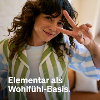 Frau mit lockigem Haar in einem gestreiften Hemd lächelt und zeigt mit ihren Fingern ein Friedenszeichen. Der Text unten lautet: „Elementar als Wohlfühl-Basis.“.