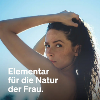 Eine Frau mit nassem Haar blickt in die Kamera, während sie ihr Haar hochhält, im Hintergrund Sonnenlicht. Deutscher Text lautet: „Elementar für die Natur der Frau.“.