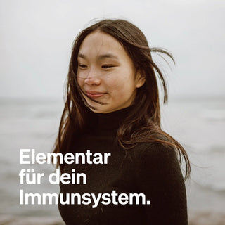 Eine junge Frau mit langen Haaren trägt einen schwarzen Rollkragenpullover. Der Text auf Deutsch lautet: "Elementar für dein Immunsystem.