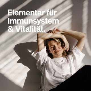 Frau in weißem Shirt sitzt, neben dem deutschen Text: "Elementar für Immunsystem & Vitalität.