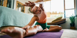 Person, die in einem Wohnzimmer auf einer Yogamatte sitzt, einen Arm über den Kopf streckt und in Richtung ihres ausgestreckten Beins greift.