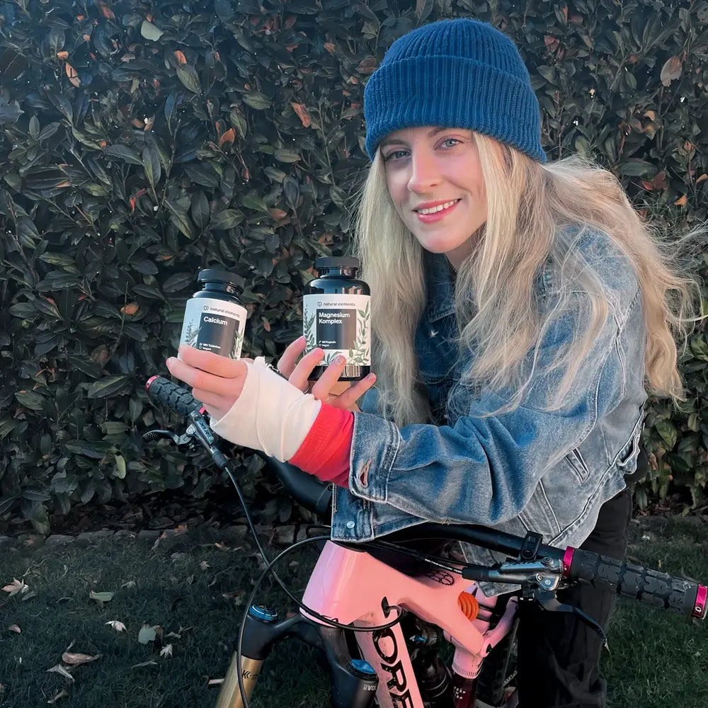 Eine blonde Frau balanciert zwei Flaschen auf ihrem Fahrrad, umgeben von Naturelementen.