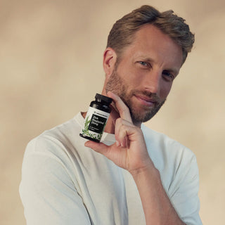 Ein Mann in einem weißen Hemd hält vor einem neutralen Hintergrund eine Flasche des Nahrungsergänzungsmittels Dr. Mercola Zinc Complex hoch.